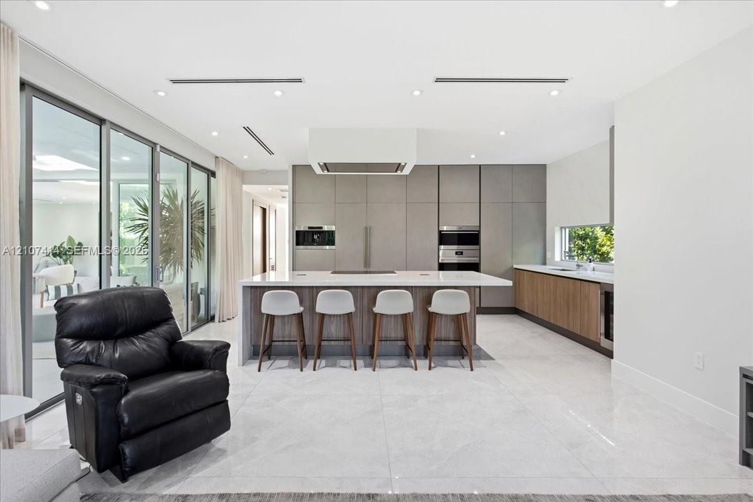 Chef’s kitchen featuring full-height matte cabinetry, a waterfall-edge stone island, built-in stainless steel appliances, polished stone flooring, and expansive floor-to-ceiling glass sliding doors