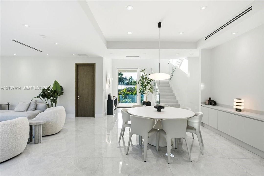Open-concept interior featuring large-format polished stone flooring, recessed LED lighting, built-in cabinetry with a stone countertop, a modern pendant light fixture, and a staircase with glass railings