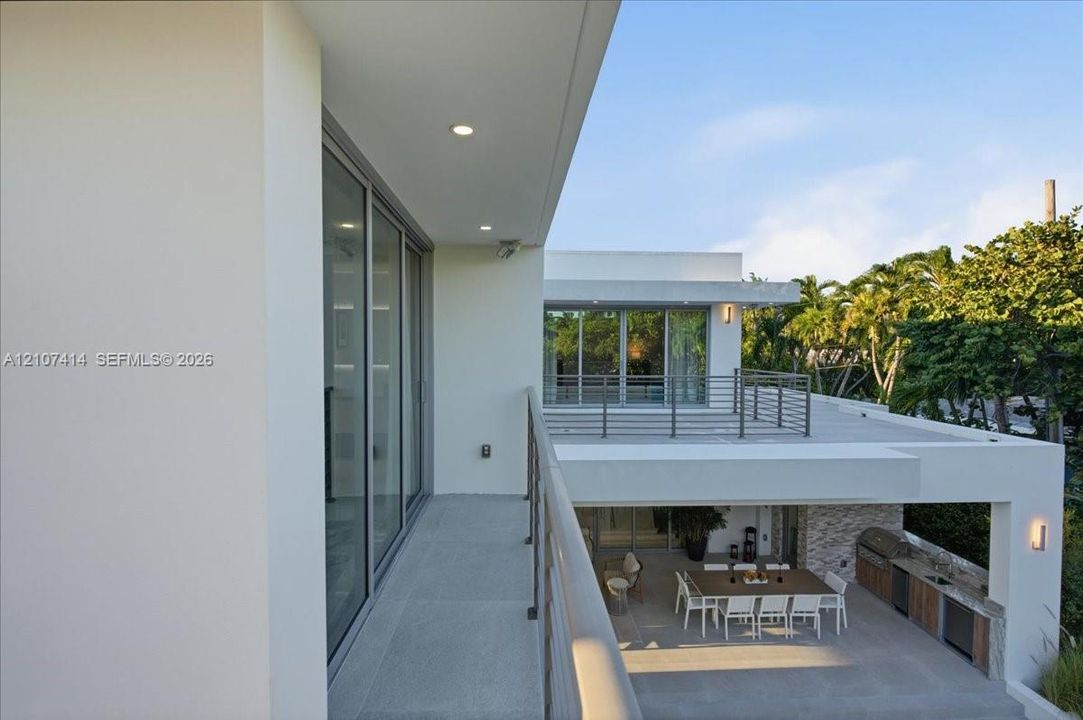 Modern architectural exterior featuring expansive covered balconies, metal railings, recessed lighting, and a ground-level outdoor kitchen with stone veneer detailing