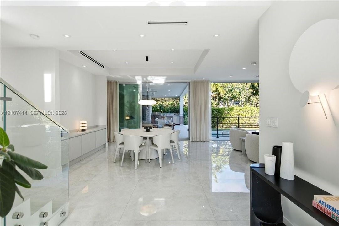 Large-format polished stone flooring, recessed ceiling lighting with tray detail, glass-panel staircase railing, built-in cabinetry, and expansive sliding glass doors