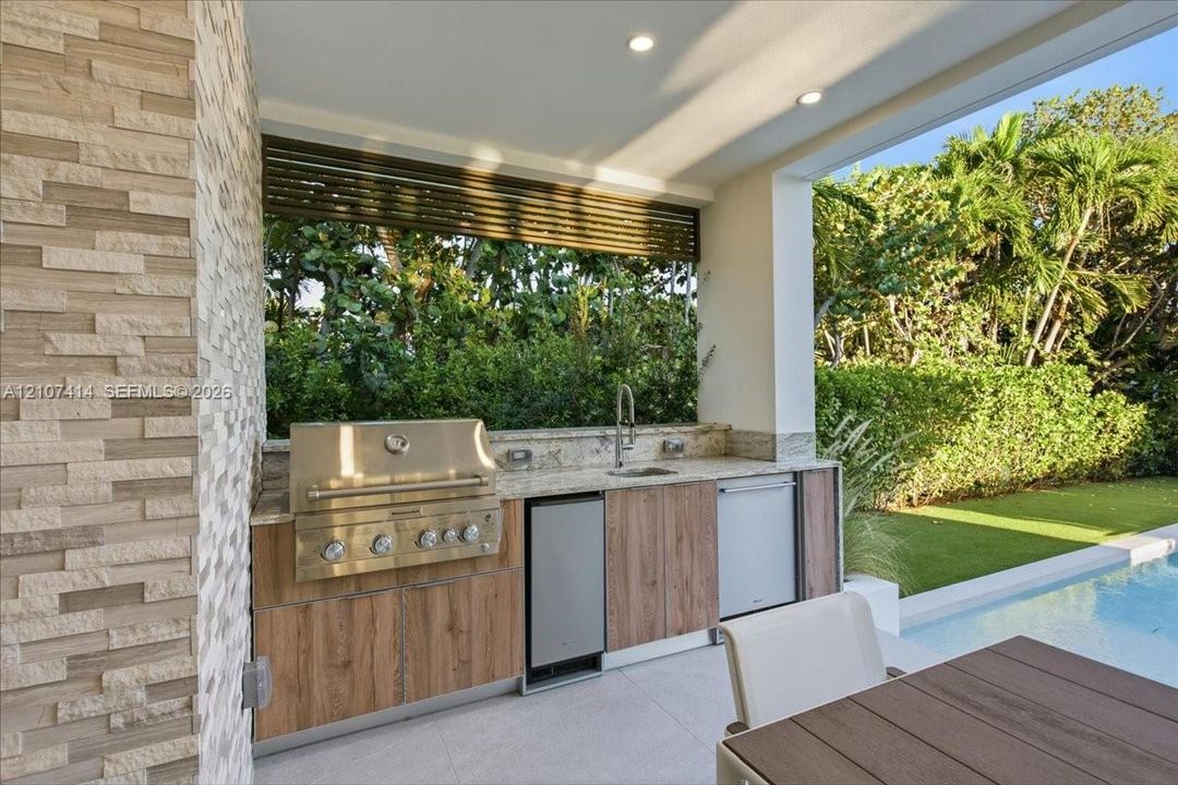 Outdoor kitchen featuring a stone-clad wall, built-in stainless steel grill, stone countertop with integrated sink, wood-finish cabinetry, and recessed lighting