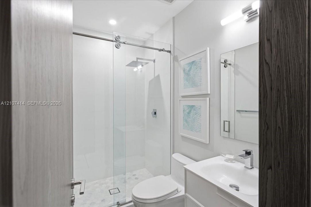 Walk-in shower with sliding glass enclosure and rainfall showerhead, white vanity with integrated basin, polished chrome fixtures, and wall-mounted vanity lighting