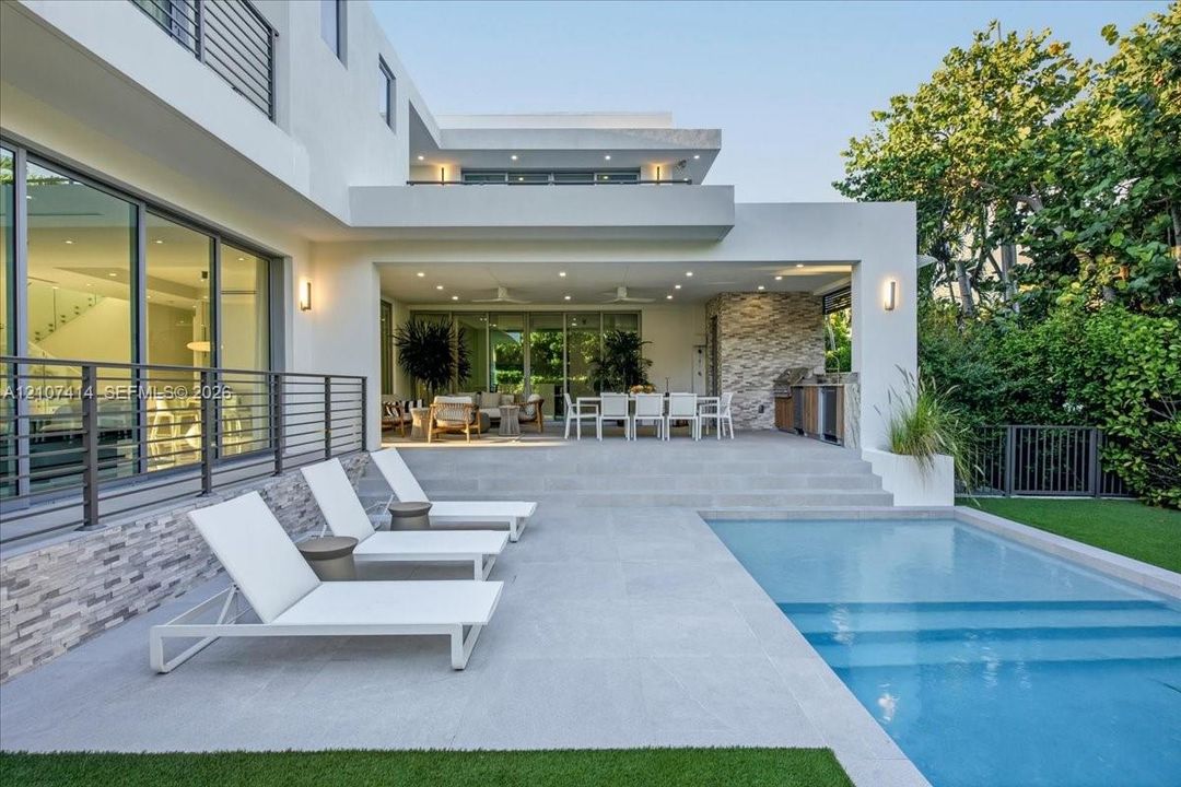 Modern multi-level exterior featuring a covered outdoor kitchen, stone accent wall, swimming pool with stepped entry, and large-format paved decking