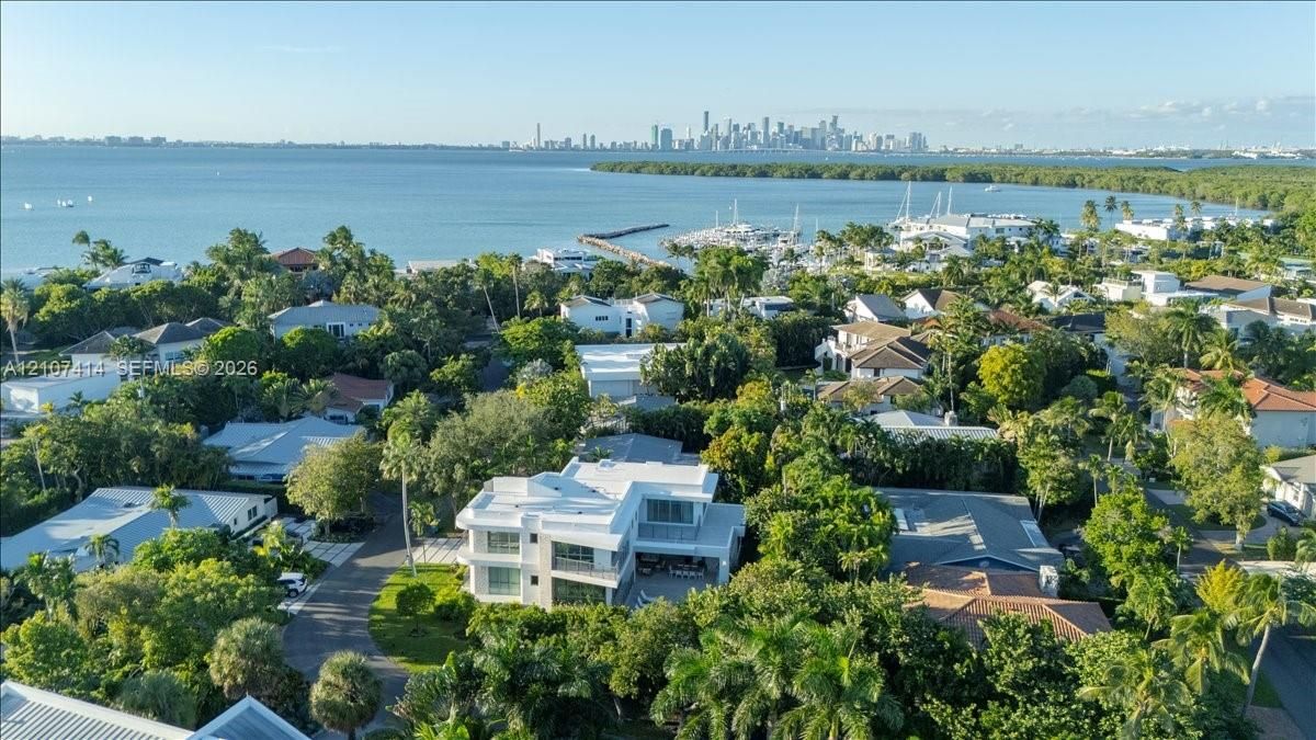 Aerial perspective featuring a two-story residence with a flat roof, large glass pane windows, and a paved driveway, set against a coastal backdrop with a prominent city skyline