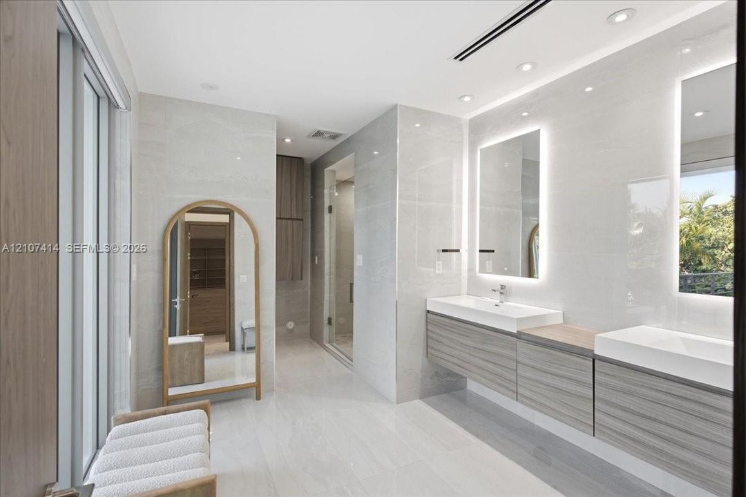 Dual floating vanity with horizontal wood-finish cabinetry, integrated rectangular sinks, and backlit mirrors, accented by large-format neutral wall tiles and recessed lighting