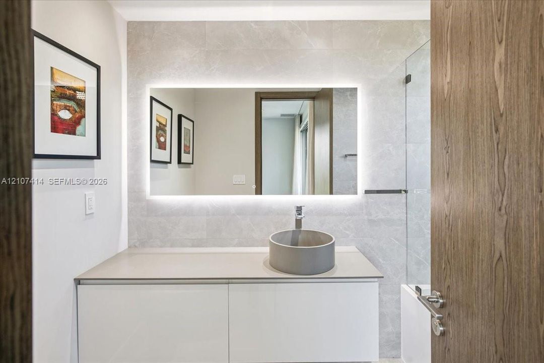 White vanity with a solid-surface countertop, cylindrical vessel sink, LED-backlit mirror, and full-height stone-finish wall tiling
