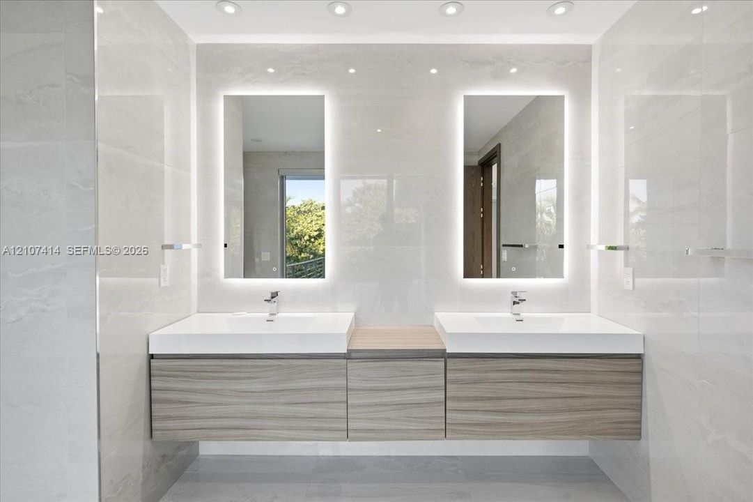 Floating dual vanity with wood-finish cabinetry, white integrated sinks, backlit rectangular mirrors, and recessed ceiling lighting, set against large-format tiled walls