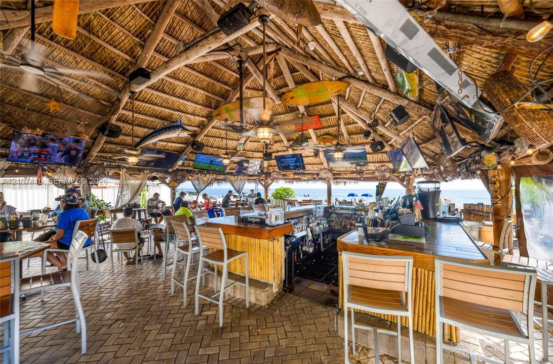 Open-air pavilion featuring a vaulted thatched roof, exposed timber framing, a central bar with bamboo-style paneling, and brick paver flooring