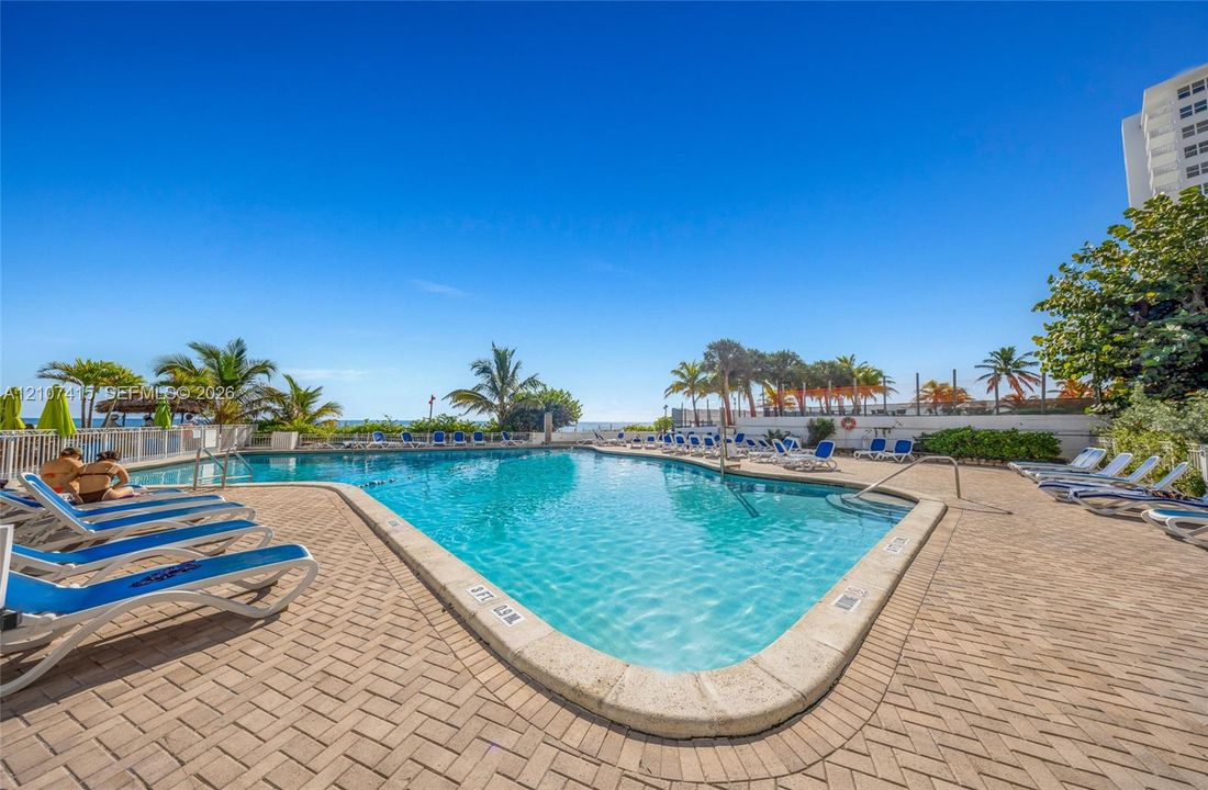 Swimming pool featuring a herringbone-patterned paver deck, metal perimeter fencing, integrated steps, and mature palm trees