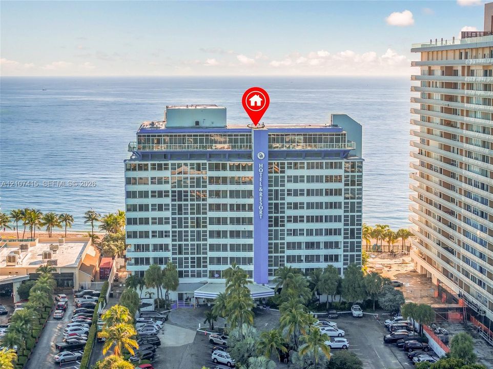 High-rise coastal building featuring floor-to-ceiling windows, an expansive glass-enclosed top level, and mature palm tree landscaping