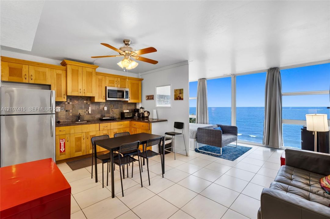 Kitchen featuring light wood cabinetry, stone tile backsplash, stainless steel refrigerator, and integrated microwave, complemented by floor-to-ceiling windows with ocean scenery and tile flooring