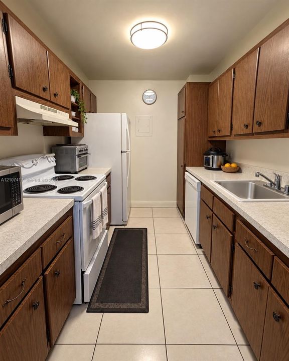 Galley-style kitchen featuring wood-finish cabinetry, solid-surface countertops, tile flooring, built-in range hood, and a ceiling-mounted light fixture