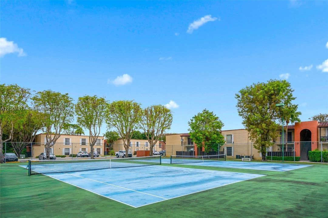 Dual tennis courts featuring hard-surface playing areas, perimeter chain-link fencing, and mature surrounding landscaping
