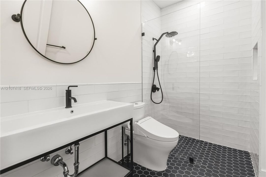 Wall-mounted round mirror, console-style vanity with a rectangular sink, matte black faucet hardware, glass-enclosed shower with subway tile surround, and hexagonal floor tiling