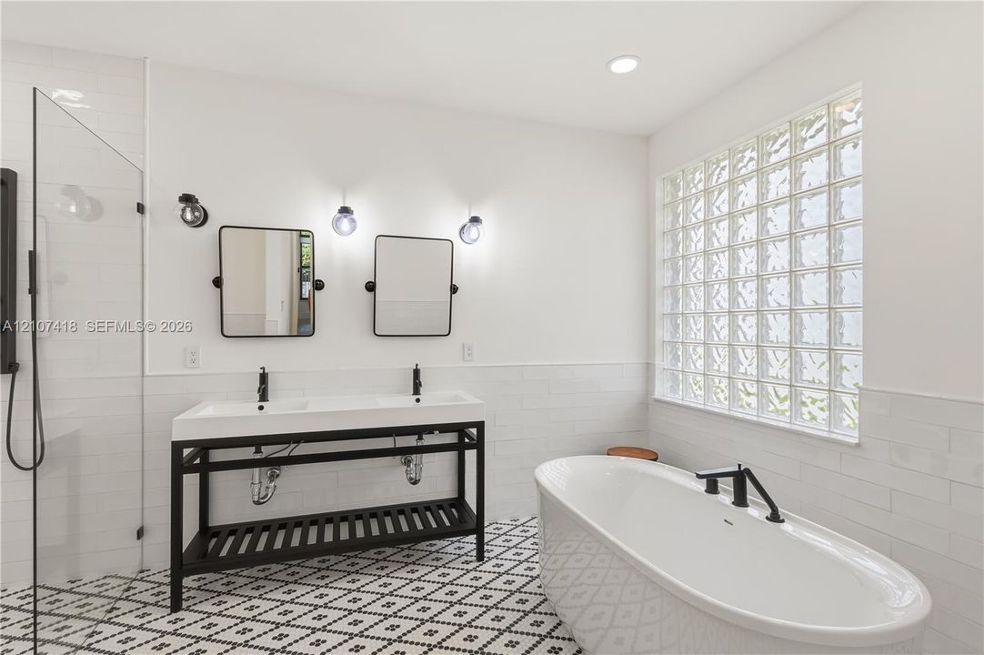 Double vanity with matte black hardware, freestanding soaking tub, walk-in glass-enclosed shower, patterned floor tile, and glass block window