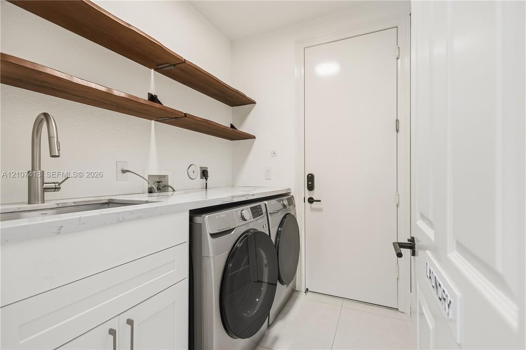 Laundry room featuring white cabinetry, stone-finish countertops, an undermount sink with a high-arch faucet, wall-mounted wood-finish shelving, and large-format tile flooring