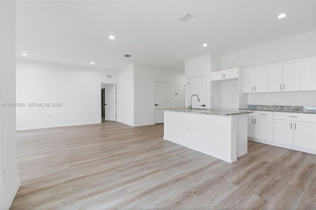 Open-concept kitchen featuring white shaker-style cabinetry, stone-surface countertops, a center island with a stainless steel pull-down faucet, and wood-finish flooring