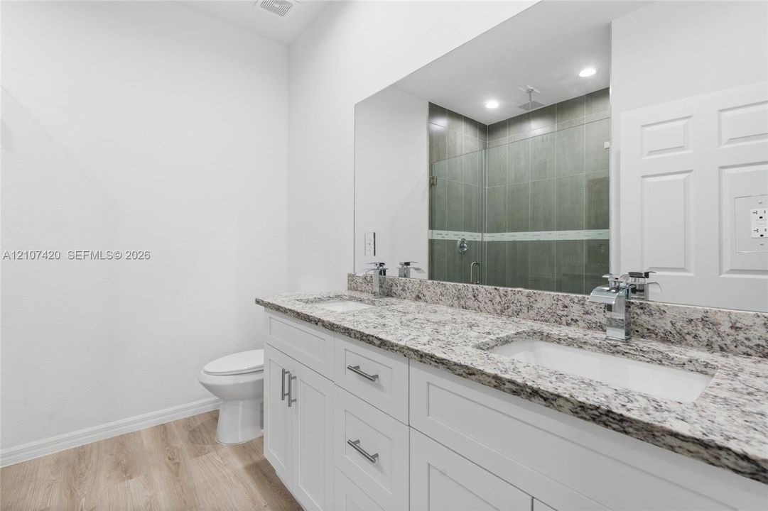 Dual-sink vanity with stone-finish countertop, white cabinetry with silver-tone hardware, wood-finish flooring, and a walk-in shower with tiled surround