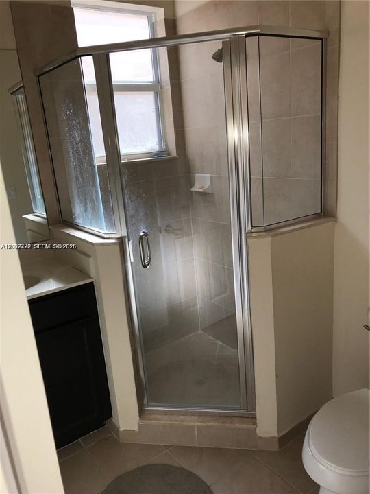 Corner shower stall with a glass enclosure, neutral tile surround, vanity with a light-toned countertop, and tile flooring
