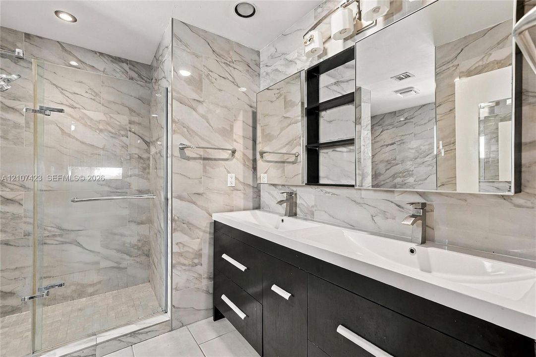 Dual-sink vanity with dark cabinetry and metallic hardware, large wall-to-wall mirror, glass-enclosed shower, and full-height wall tile
