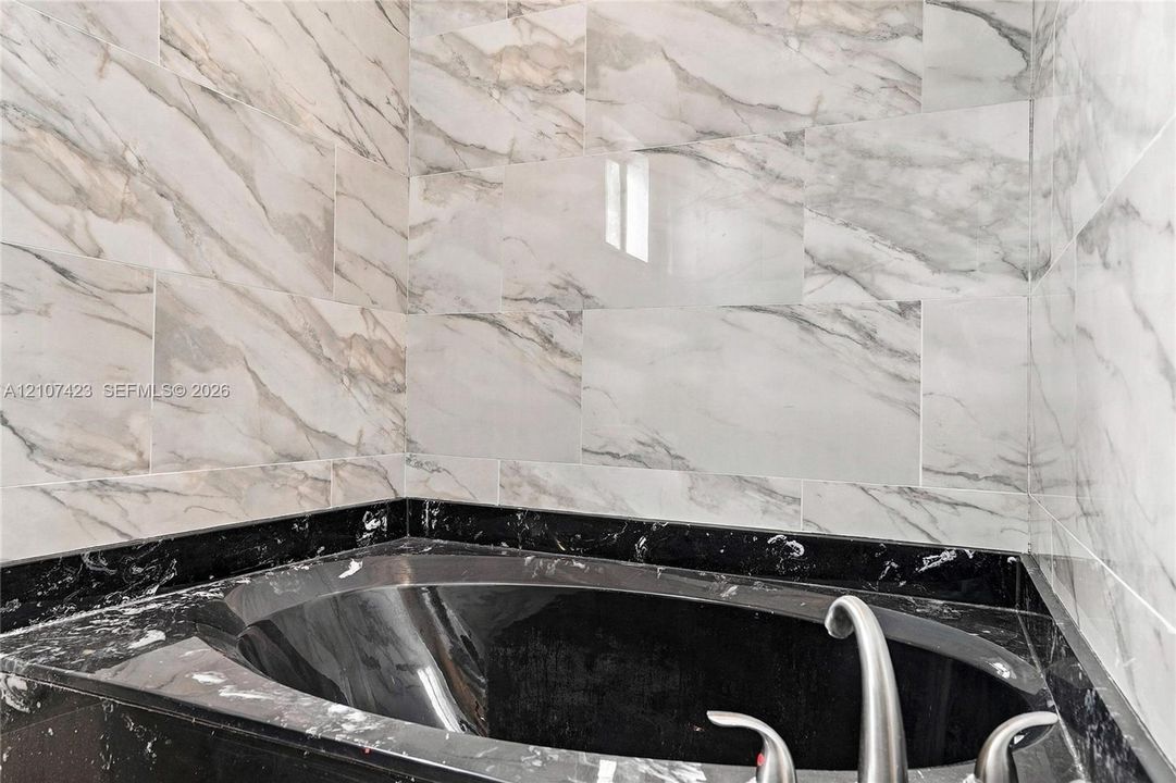 Built-in soaking tub with a dark stone-finish surround, complemented by floor-to-ceiling wall tile featuring a veined pattern and a metallic faucet fixture
