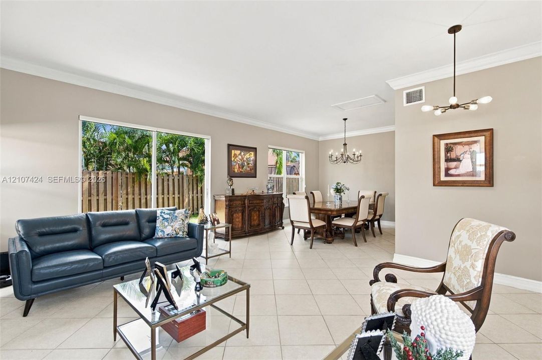Open-concept living space featuring large-format ceramic tile flooring, neutral wall tones, crown molding, sliding glass doors, and two multi-arm chandeliers