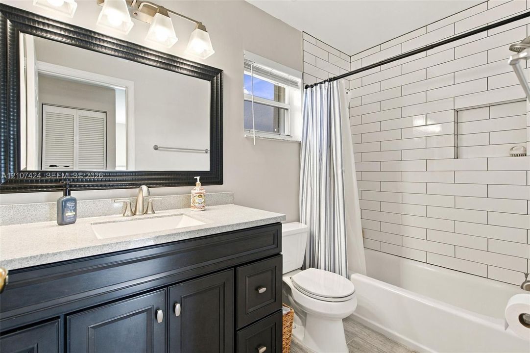 Bathroom featuring a dark-toned vanity with a light stone-surface countertop, rectangular undermount sink, framed wall mirror with integrated multi-bulb lighting, and a shower-tub combination finished with horizontal subway tiling and a recessed niche