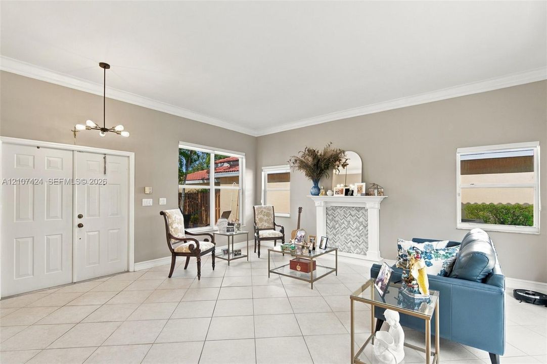 Open living area featuring large-format diagonal tile flooring, crown molding, double-door entryway, decorative fireplace with mosaic tile surround, and multi-light chandelier