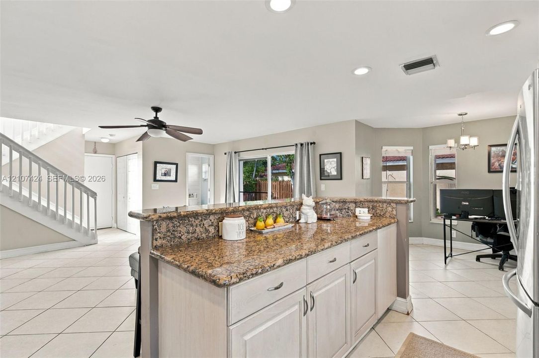 Kitchen featuring a stone-topped island with shaker-style cabinetry, recessed lighting, neutral ceramic tile flooring, and a staircase with a wooden handrail