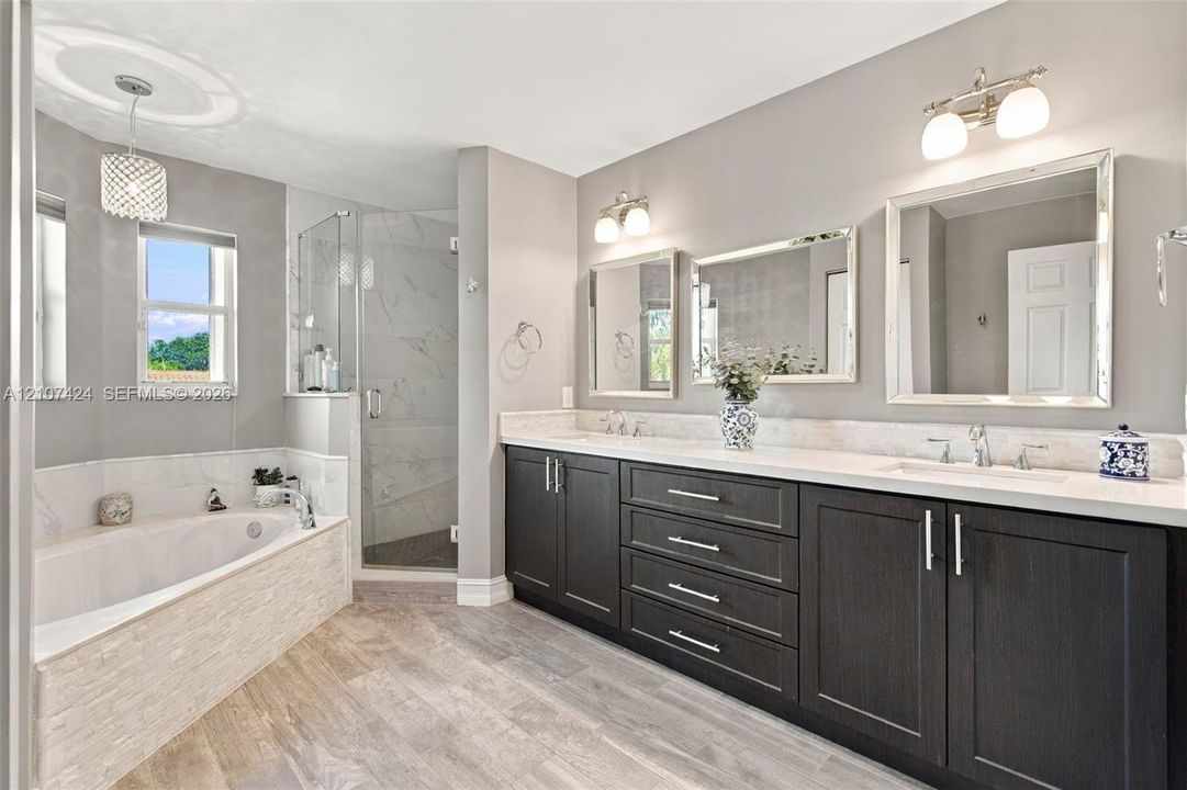 Dual-sink vanity with dark cabinetry, light-toned countertop, and framed mirrors, featuring a separate soaking tub with stone-textured surround and a glass-enclosed shower with tiled interior