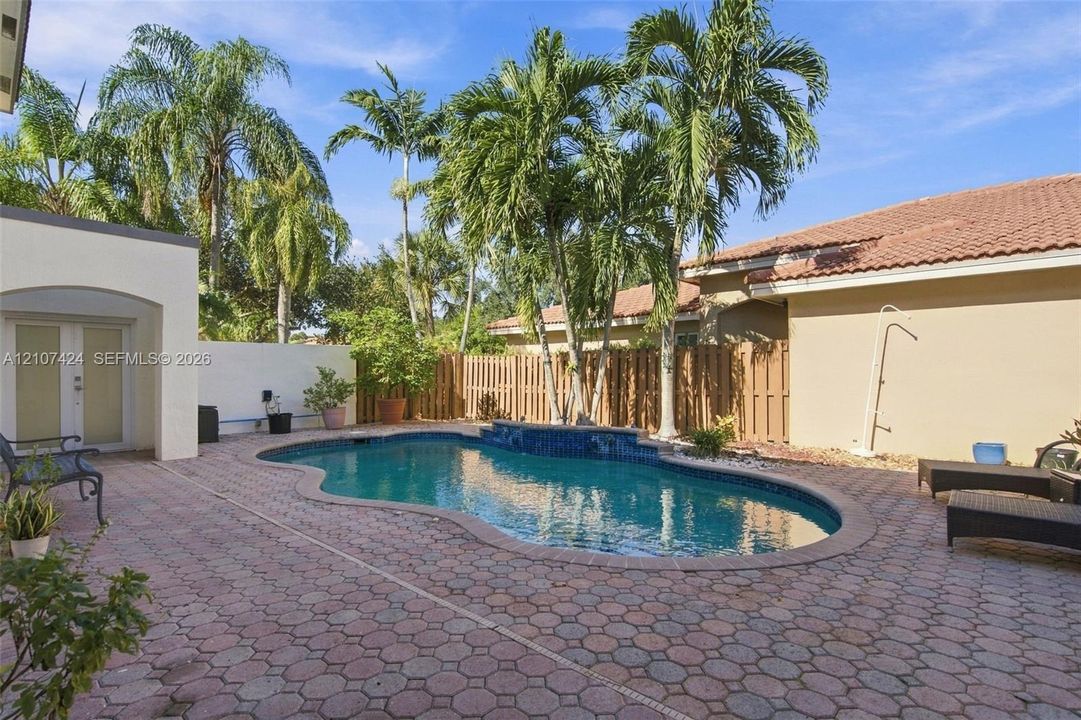 Outdoor swimming pool featuring a decorative tile water feature, surrounded by extensive patterned paver hardscaping, mature palm trees, and full-height privacy fencing