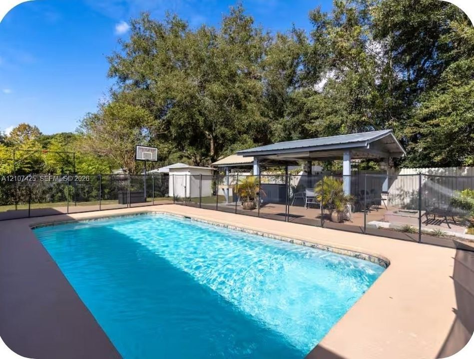 In-ground swimming pool with surrounding concrete decking, mesh safety fence, metal-roofed cabana, and basketball hoop