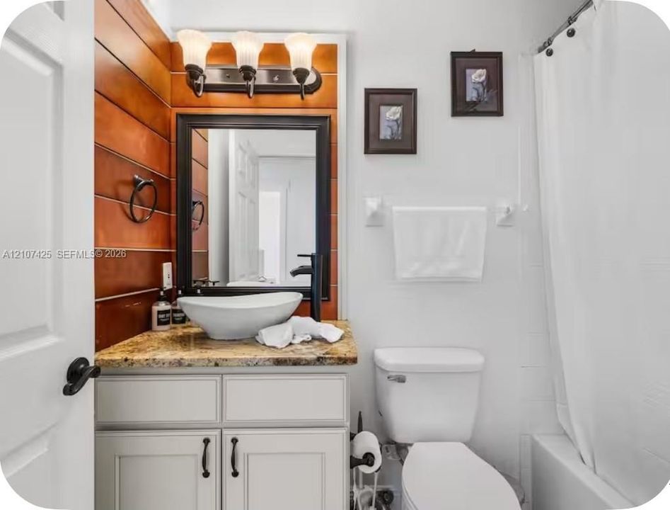 Neutral-toned vanity with a stone countertop, vessel sink, matte black hardware, and accent wall with wood-finish paneling