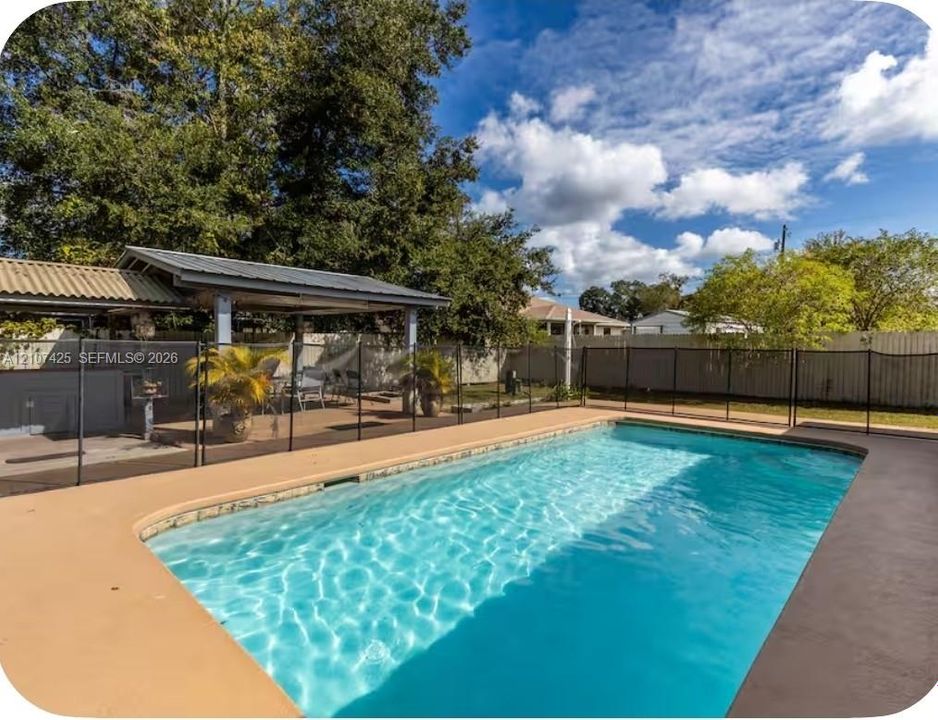 In-ground swimming pool with surrounding concrete decking, mesh safety fencing, and a covered outdoor patio area