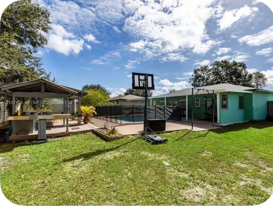 Expansive backyard featuring a covered outdoor kitchen, concrete patio, swimming pool with safety fencing, and an open green lawn