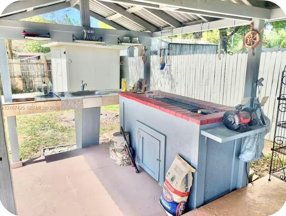 Covered outdoor kitchen featuring a built-in barbecue grill with brick surround, stainless steel sink, tile-topped prep counters, and structural wooden framing