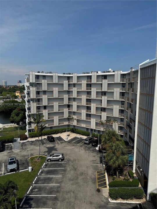 Mid-rise residential building featuring a structural geometric facade, stacked private balconies, dedicated surface parking, and mature palm landscaping