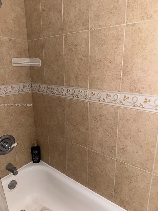 Bathtub surround featuring neutral-toned square wall tiles, a decorative floral border inlay, integrated corner soap dish, and chrome-finish plumbing fixtures