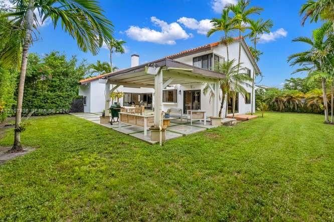 Rear exterior featuring a covered patio with concrete pavers, a wooden pergola, and a manicured lawn framed by mature palm trees