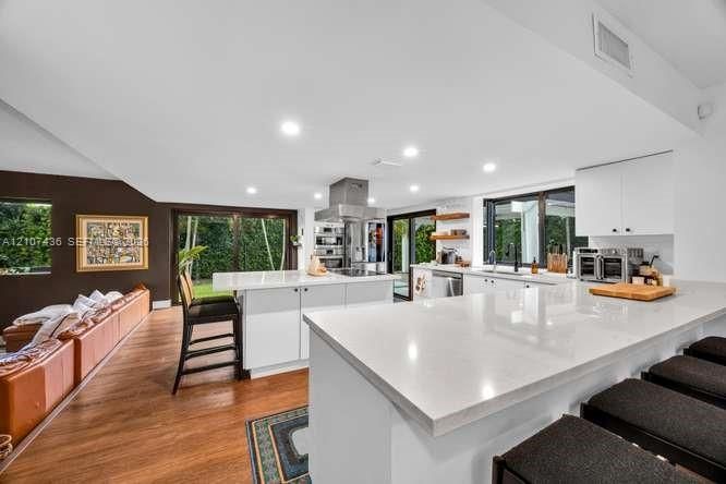 Modern kitchen featuring an expansive central island with quartz countertops, custom white cabinetry, stainless steel range hood, recessed lighting, and wood-finish flooring