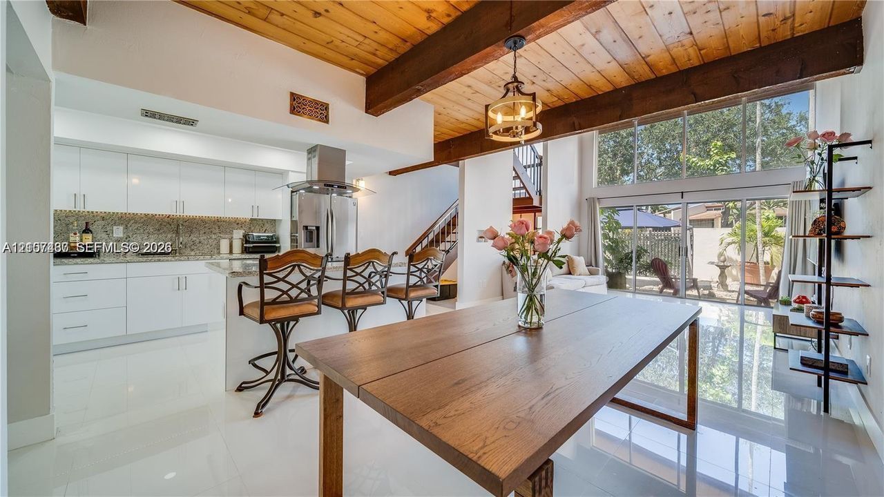 Open-concept kitchen and dining area featuring vaulted ceilings with exposed wood beams, white cabinetry, stone countertops, and full-height glass windows