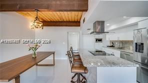 Open-concept kitchen featuring an oversized stone-topped island with stainless steel appliances, exposed wood-beam ceiling, wood-finish dining surface, and hanging decorative lighting fixture