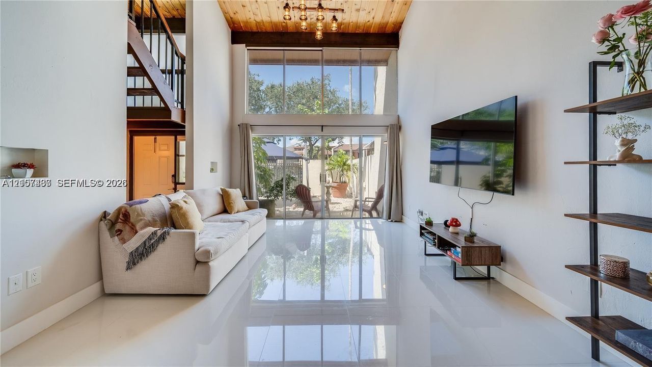 Double-height living area featuring vaulted wood-plank ceiling, exposed wood beams, polished porcelain tile flooring, and floor-to-ceiling glass sliding doors