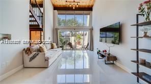 Double-height living area featuring floor-to-ceiling glass windows, high wood-beam ceilings, polished tile flooring, and an open-concept mezzanine staircase
