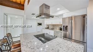 Kitchen featuring a stone-topped island with an integrated cooktop, stainless steel refrigerator, professional-grade range hood, white cabinetry, and exposed wood ceiling beam