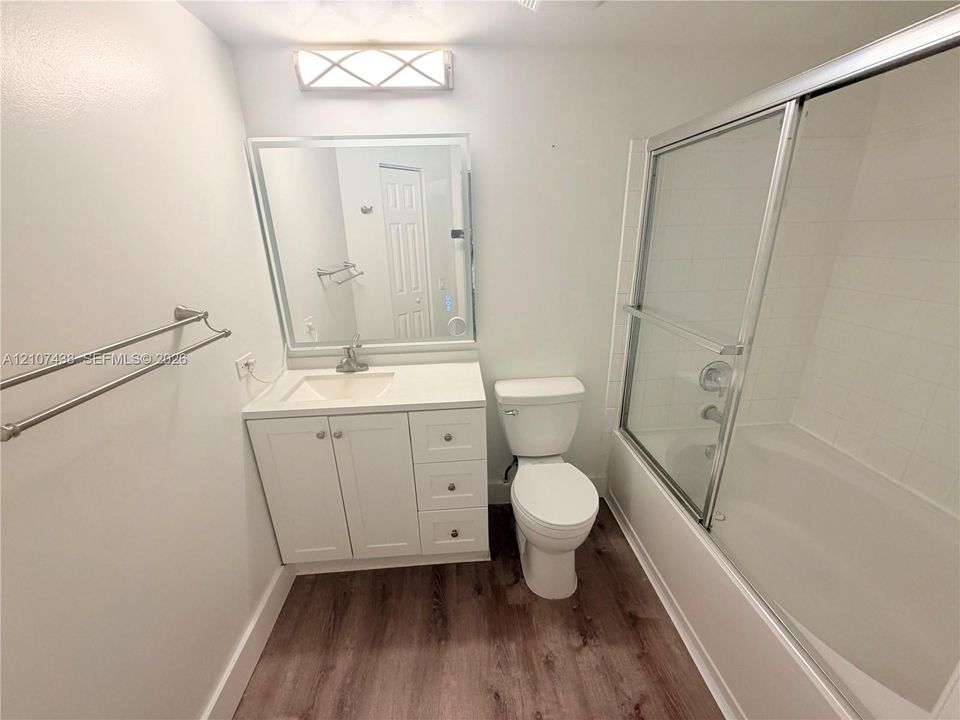 Bathroom featuring wood-finish flooring, white vanity with drawers, rectangular mirror, glass-enclosed tub shower with tiled surround, and integrated lighting