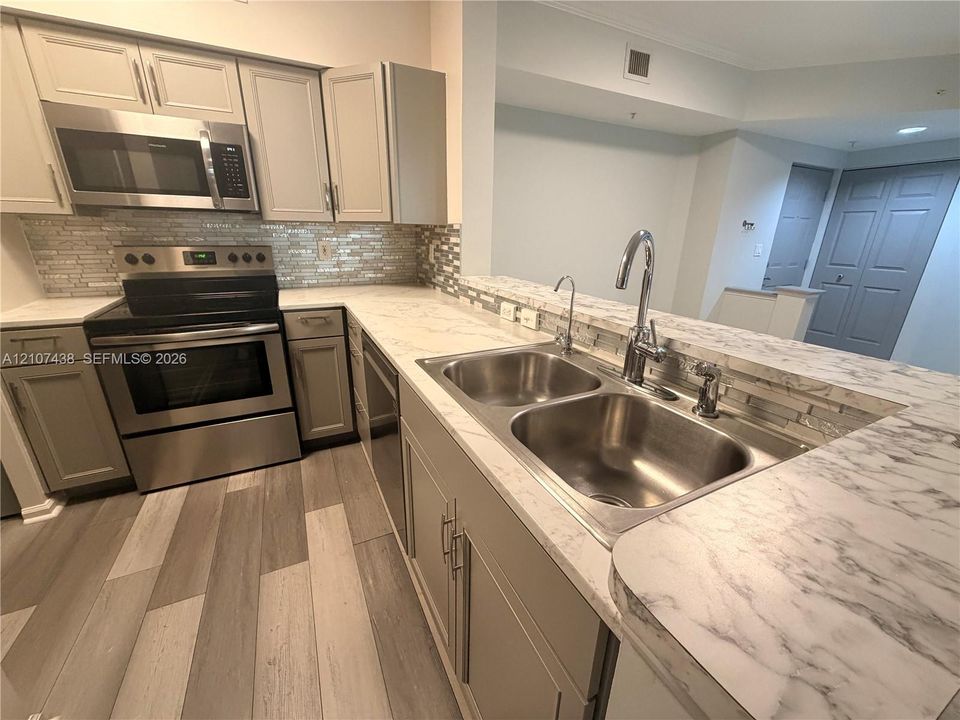 Kitchen featuring light-toned cabinetry, stainless steel appliances, stone-patterned countertops, a dual-basin stainless steel sink with a high-arch faucet, and wood-finish flooring