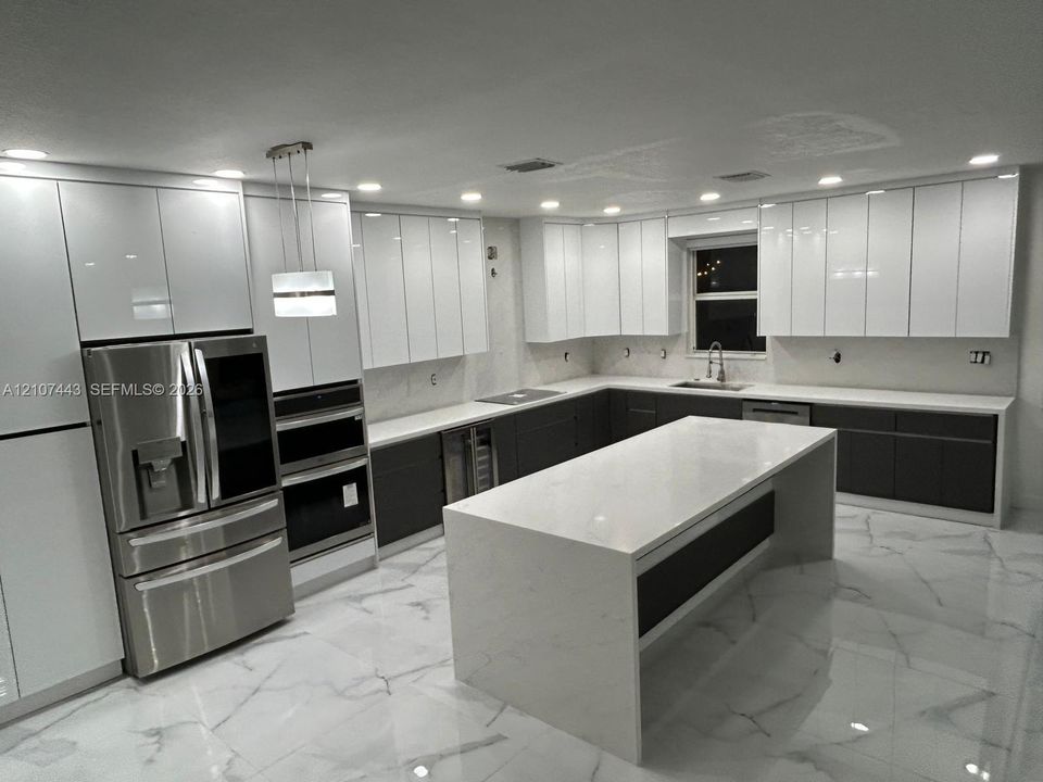 Kitchen featuring high-gloss white upper cabinetry, dark lower cabinetry, a large central island with a stone-finish waterfall countertop, stainless steel appliances, and large-format tile flooring