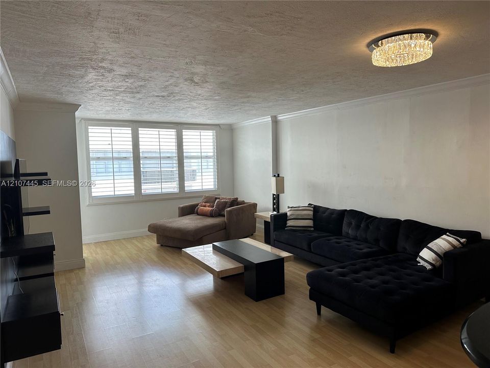 Living area featuring wood-finish flooring, plantation shutters, crown molding, and a crystal-accented flush mount ceiling light