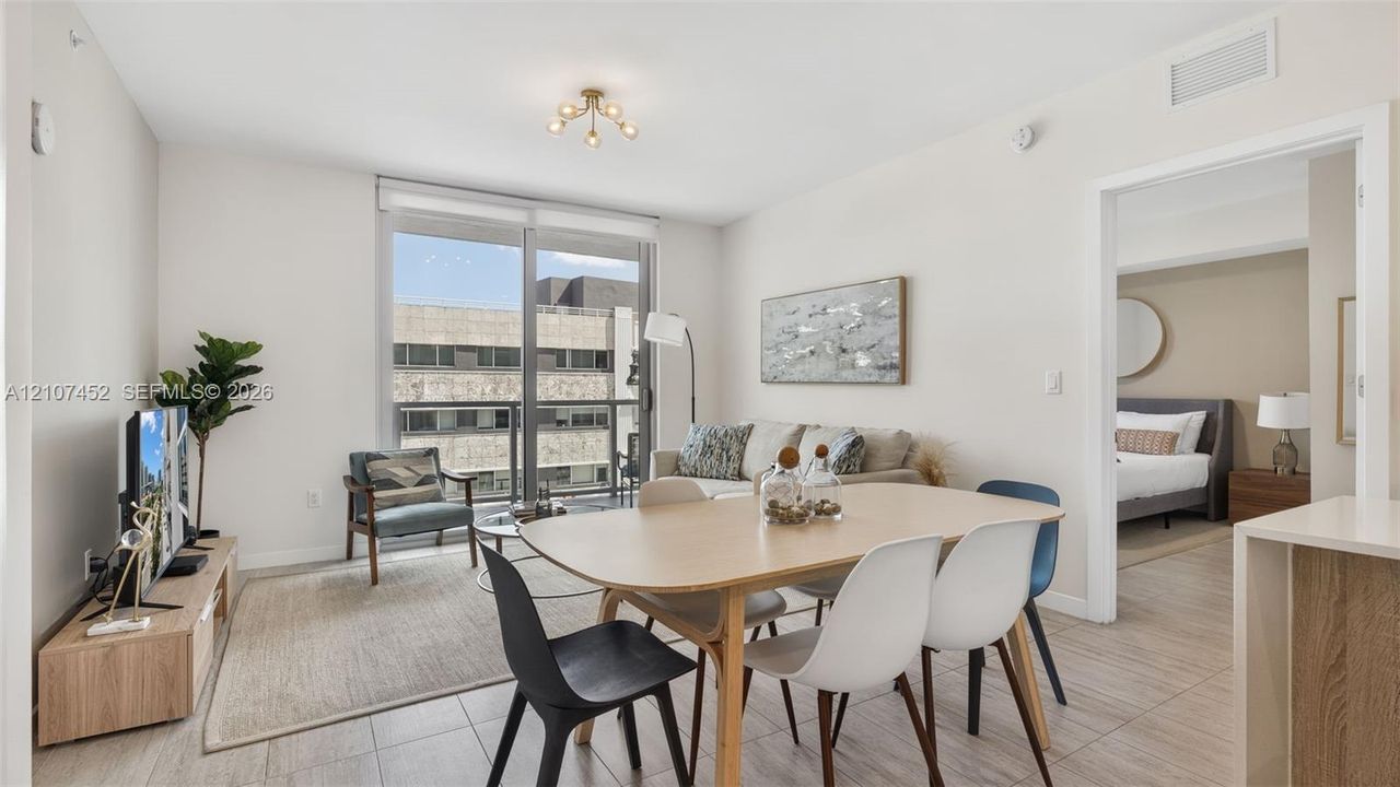 Open-concept interior featuring large-format ceramic tile flooring, neutral wall tones, multi-light flush-mount ceiling fixture, and full-height sliding glass doors leading to a private balcony
